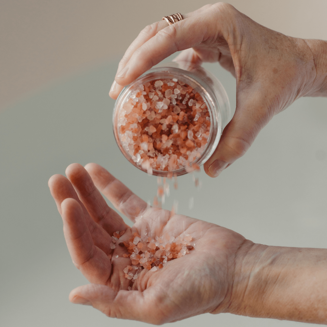 A person pours Palermo Body’s Replenishing Salt Soak from a glass jar into their palm, pink Himalayan salt crystals spilling gently from hand to hand.