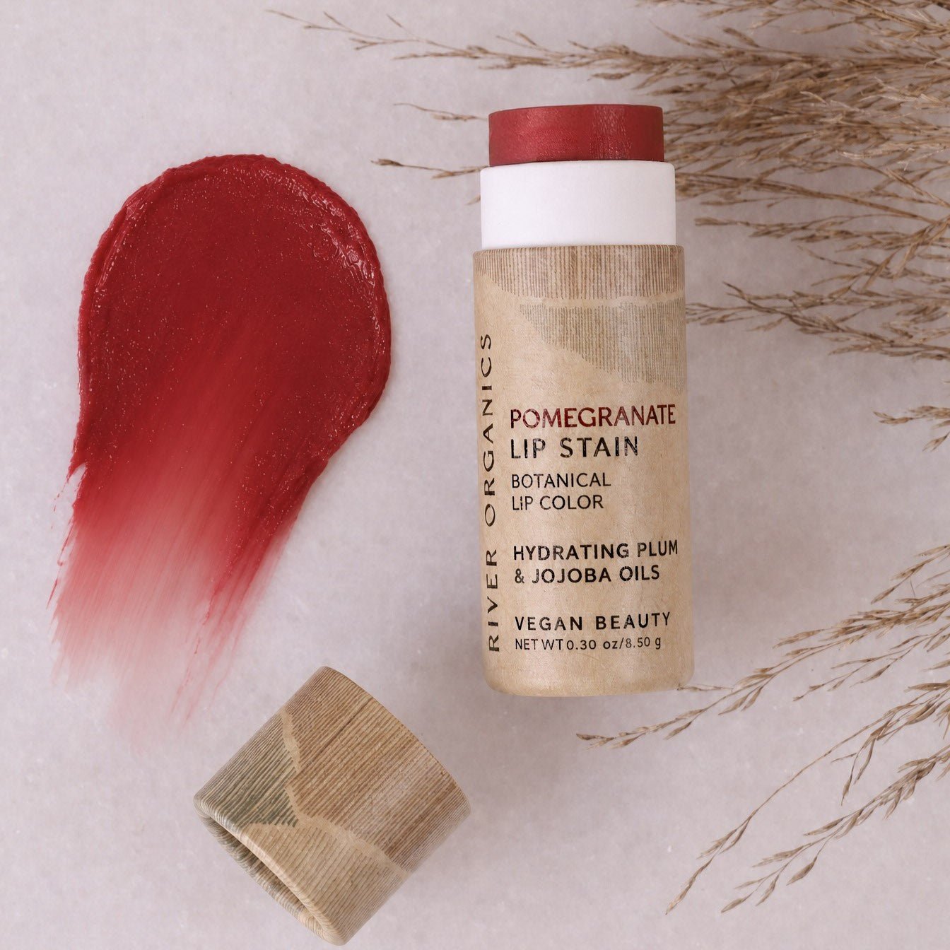 A tube of River Organics Lip Stain: Vegan, Plastic-Free, & Organic | All Colors with its cap off sits on a marble surface adorned with dried grass, its vibrant red pigment smeared beside it. This plastic-free and vegan formula provides a gorgeous pop of color while keeping your lips hydrated for hours.