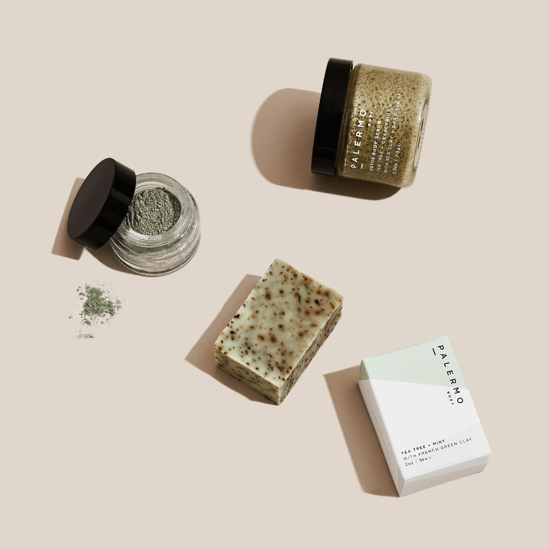 Flat lay of Palermo Body’s Detox + Clarify Mindful Kit on a beige background, featuring a green powder clay mask jar, tea tree soap bar, labeled box, and Detox body scrub with black lid.