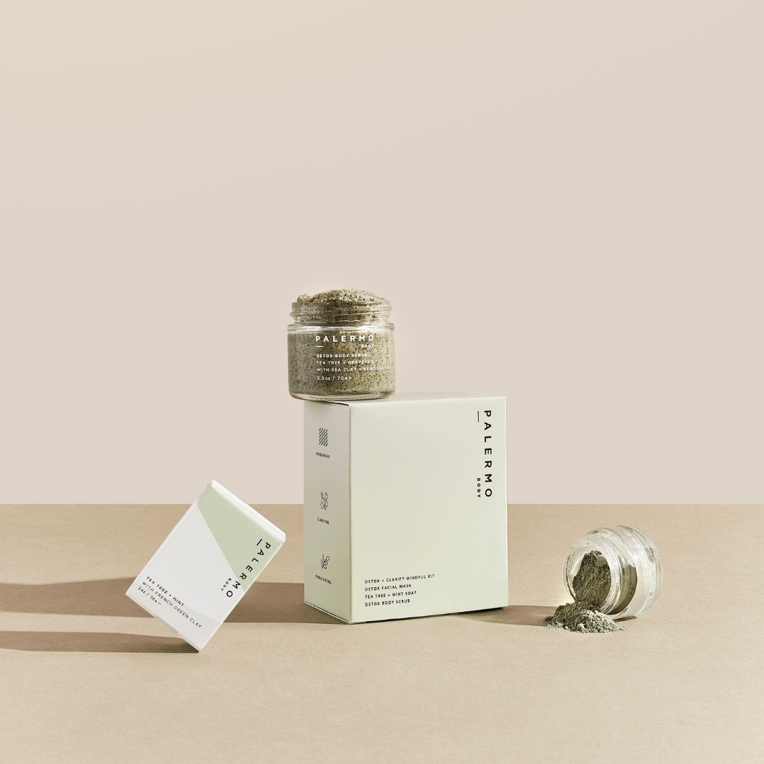 Three Palermo Body Detox + Clarify Mindful Kit containers are arranged on a beige surface, including two glass jars with green powder and two minimalist white boxes, one standing upright and one placed at an angle.