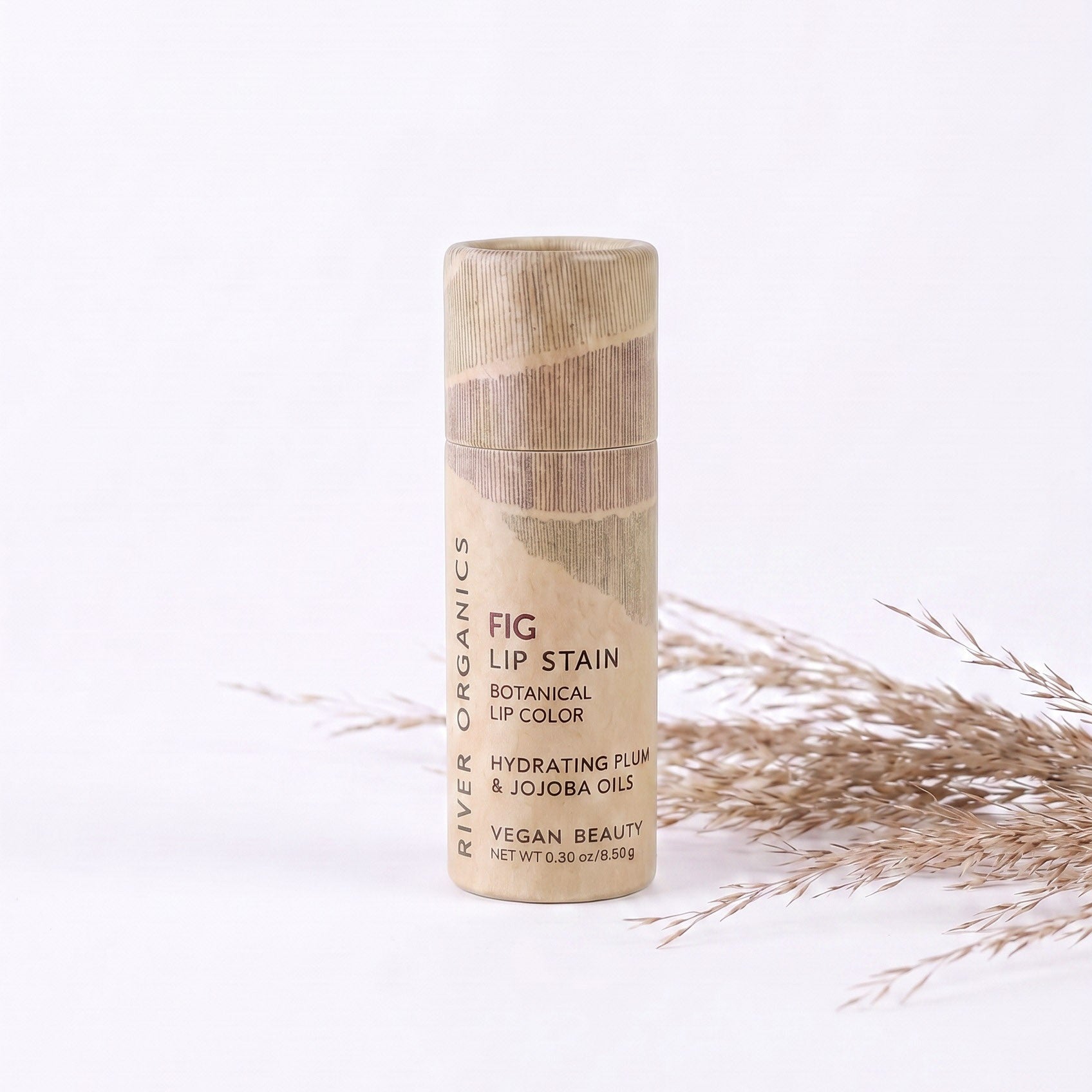 A tube of River Organics Lip Stain: Vegan, Plastic-Free, & Organic | All Colors stands upright on a white surface beside dried grass. The beige tube features brown text showcasing its vegan and botanical ingredients.