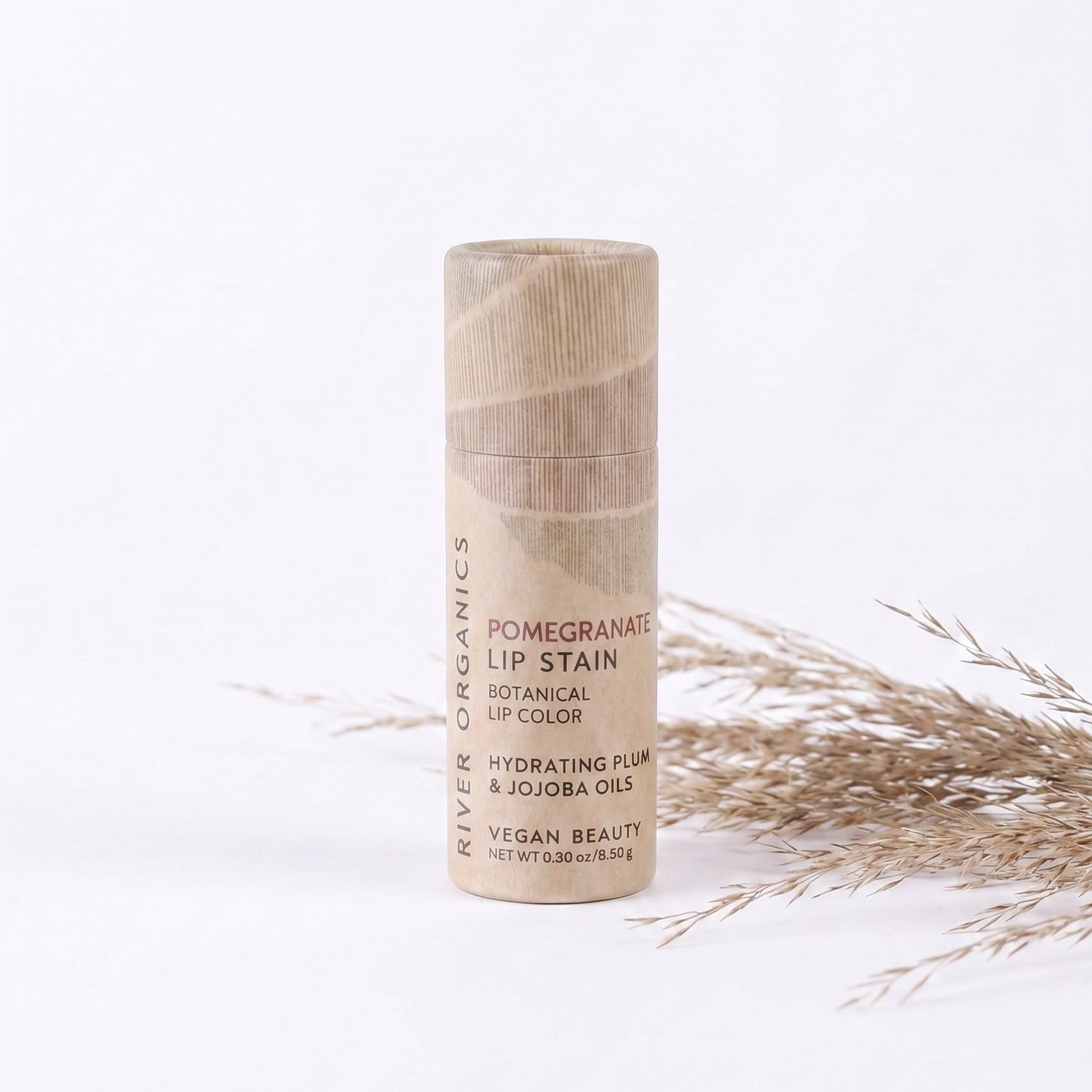 An upright tube of River Organics Lip Stain: Vegan, Plastic-Free, & Organic | All Colors is displayed on a light background, accompanied by dry decorative grass. The beige packaging features minimalist text and design, highlighting the brand’s dedication to offering a plastic-free, vegan lip stain that provides long-lasting hydration and color.