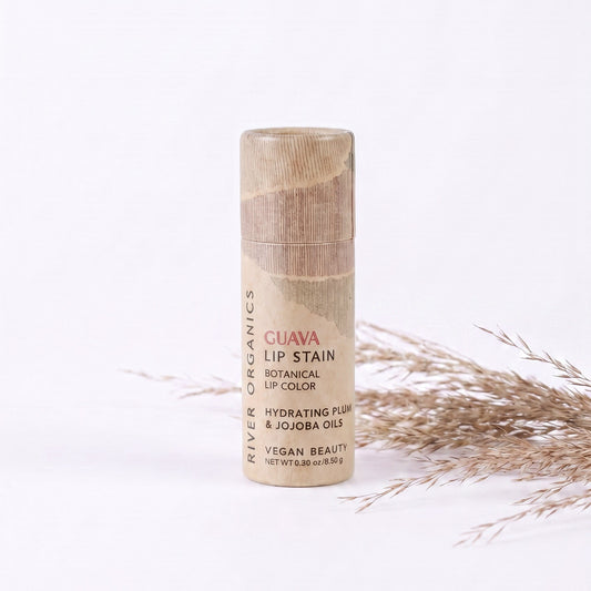A tube of River Organics Lip Stain: Vegan, Plastic-Free, & Organic | All Colors stands upright on a white surface with dried grasses in the background. The packaging highlights hydrating plum and jojoba oils and notes that it is a long-lasting, hydrating, plastic-free vegan lip stain.