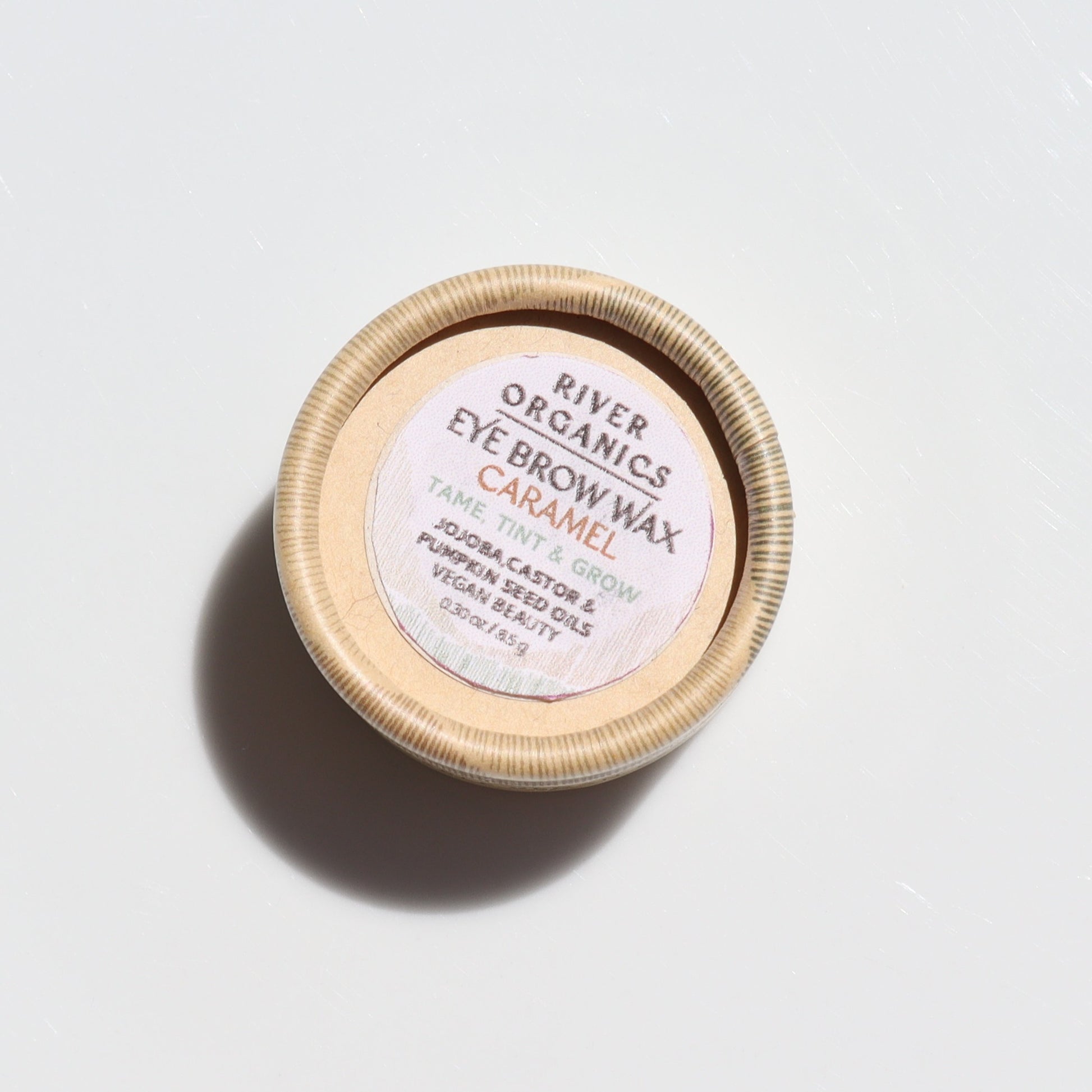 A small round wooden container of River Organics Brow Wax - Tame, Tint & Grow sits on a white surface. The label notes its vegan, organic ingredients like castor oil and beetroot for sustainable brow taming and tinting.