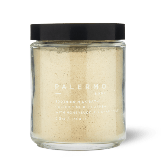 Palermo Body Soothing Milk Bath is a 5.3 oz clear glass jar with a black lid, containing light beige powder made with coconut milk, oatmeal, honeysuckle, and chamomile for soothing skin care.