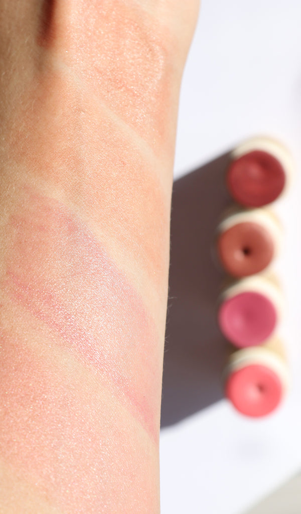 A close-up of a forearm with four swatches of River Organics Natural Cream Blush Stick | All Colors in shimmery pink and rose shades, shown with four open plastic-free containers on a white surface.
