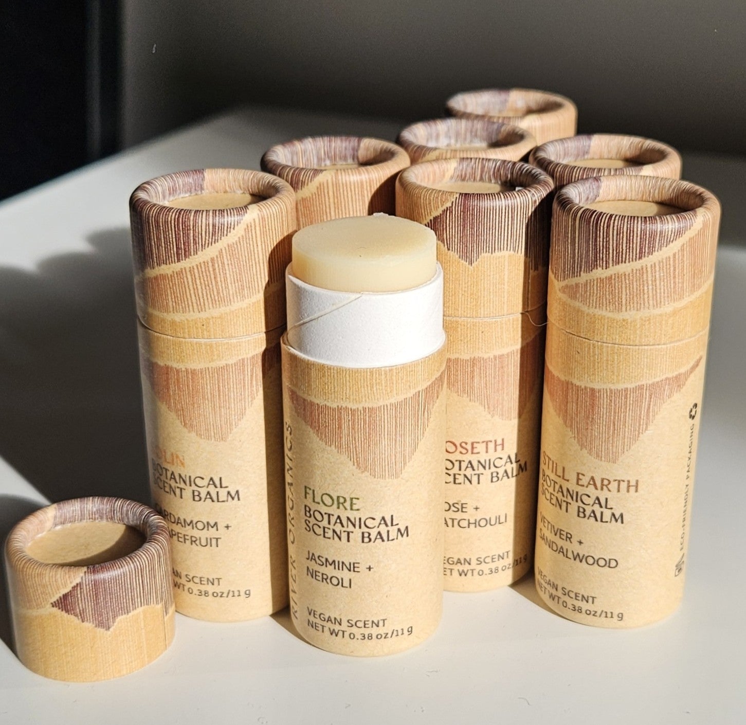 River Organics' Set of 2, 3 or 4 Botanical Scent Balms features cylindrical tubes with unique essential oil blends. One "Flore" tube is open, showing the balm inside. All labels highlight natural, vegan ingredients.