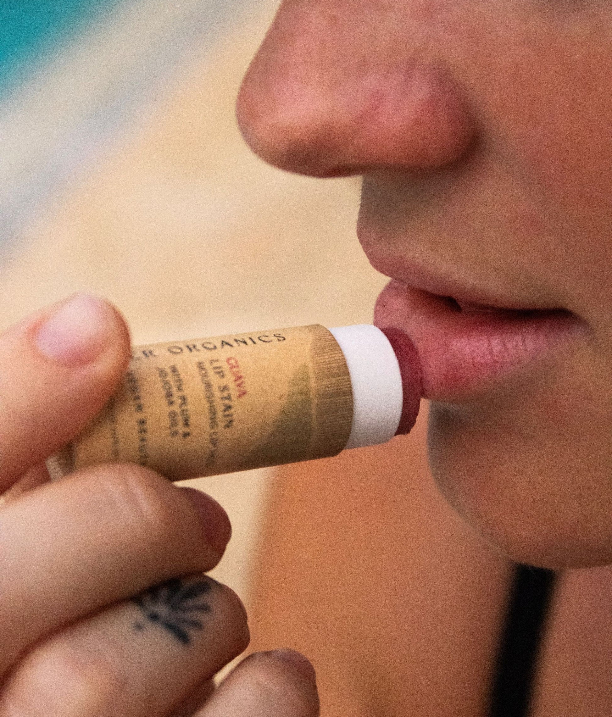 A person applies River Organics Lip Stain Sets to their lips. The close-up highlights the nose, lips, and a tattooed finger holding the lip stain.