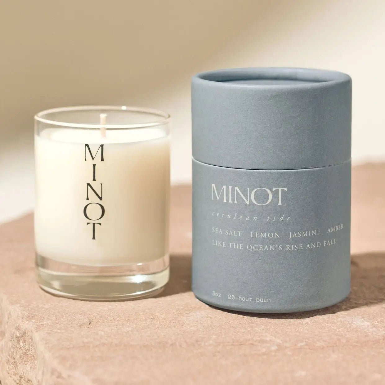 A 3oz Minot Luxury Non-toxic Soy Candle in a white glass jar sits beside a blue cylindrical box, both labeled "Minot," featuring notes of sea salt, lemon, jasmine, amber, and offering a 20-hour burn time.