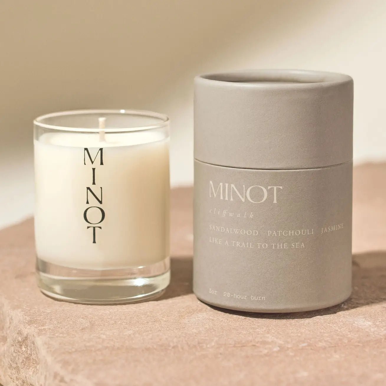 A Minot Luxury Non-toxic Soy Candle (3oz) in white patchouli scent sits in a clear glass holder labeled "MINOT," next to a gray cylindrical package reading "MINOT - SANDALWOOD PATCHOULI JASMINE" and "LIKE A TRAIL TO THE SEA" on a beige surface.
