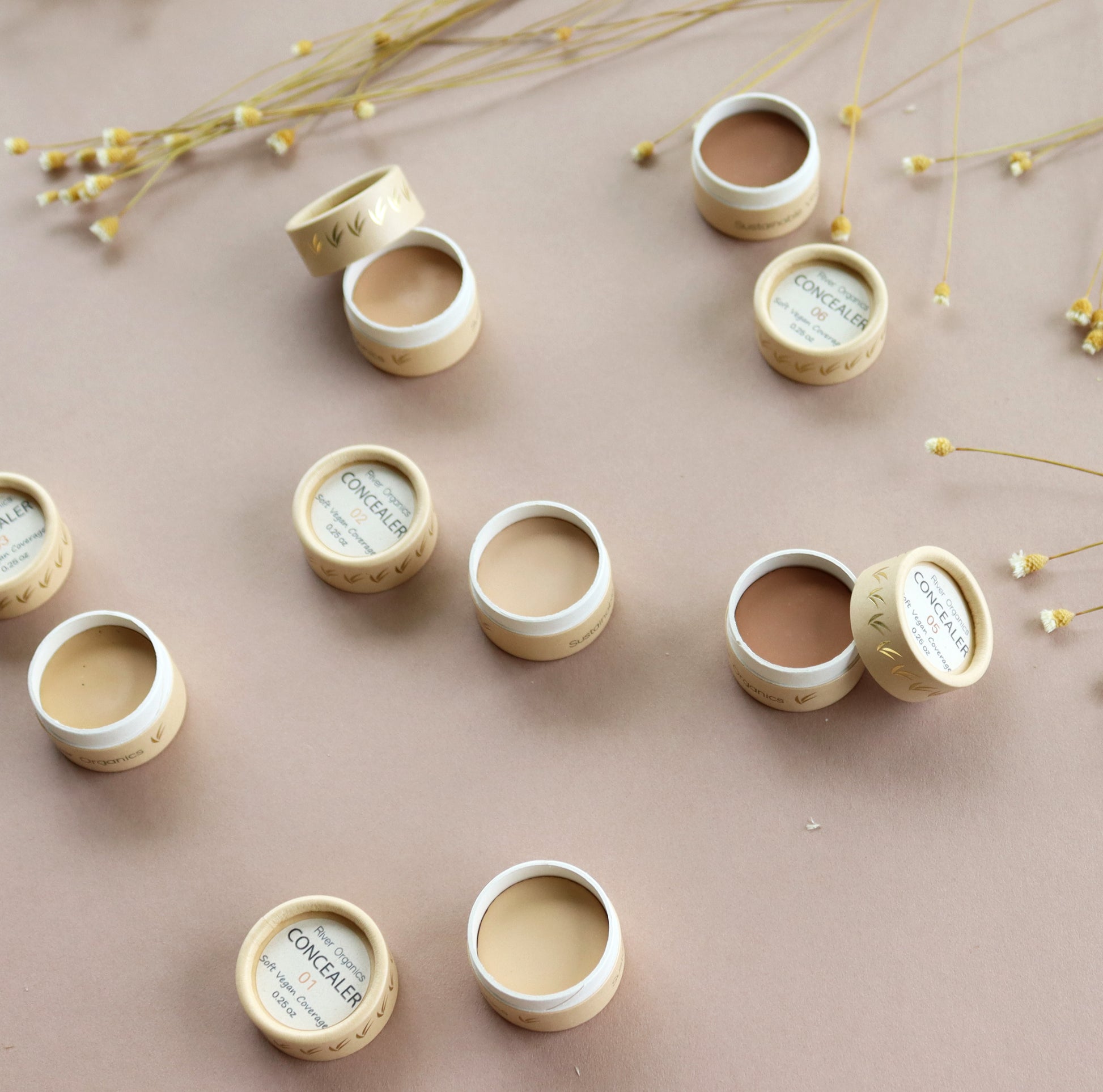 River Organics’ Zero-Waste Vegan Concealer in small round containers, displayed open in various shades on a beige surface, is accented with delicate dried flowers.