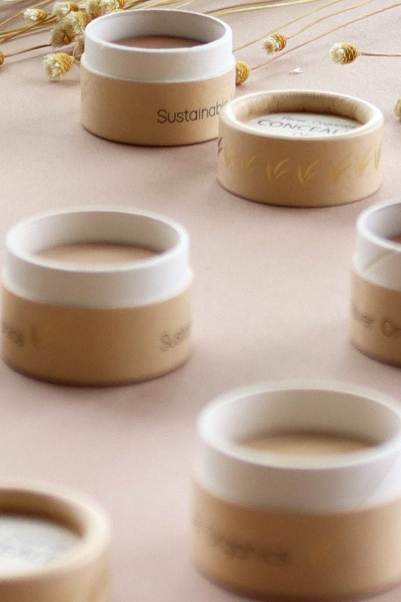 Small round containers from River Organics, featuring beige and white lids, are arranged on a soft pink surface with dried flowers scattered in the background. The labels read “Concealer & Brow Duo - Zero-Waste Vegan Beauty Set,” emphasizing the sustainable and eco-friendly focus of these beauty products.
