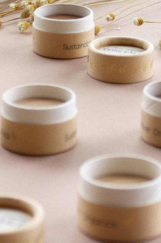 Small round containers from River Organics, featuring beige and white lids, are arranged on a soft pink surface with dried flowers scattered in the background. The labels read “Concealer & Brow Duo - Zero-Waste Vegan Beauty Set,” emphasizing the sustainable and eco-friendly focus of these beauty products.