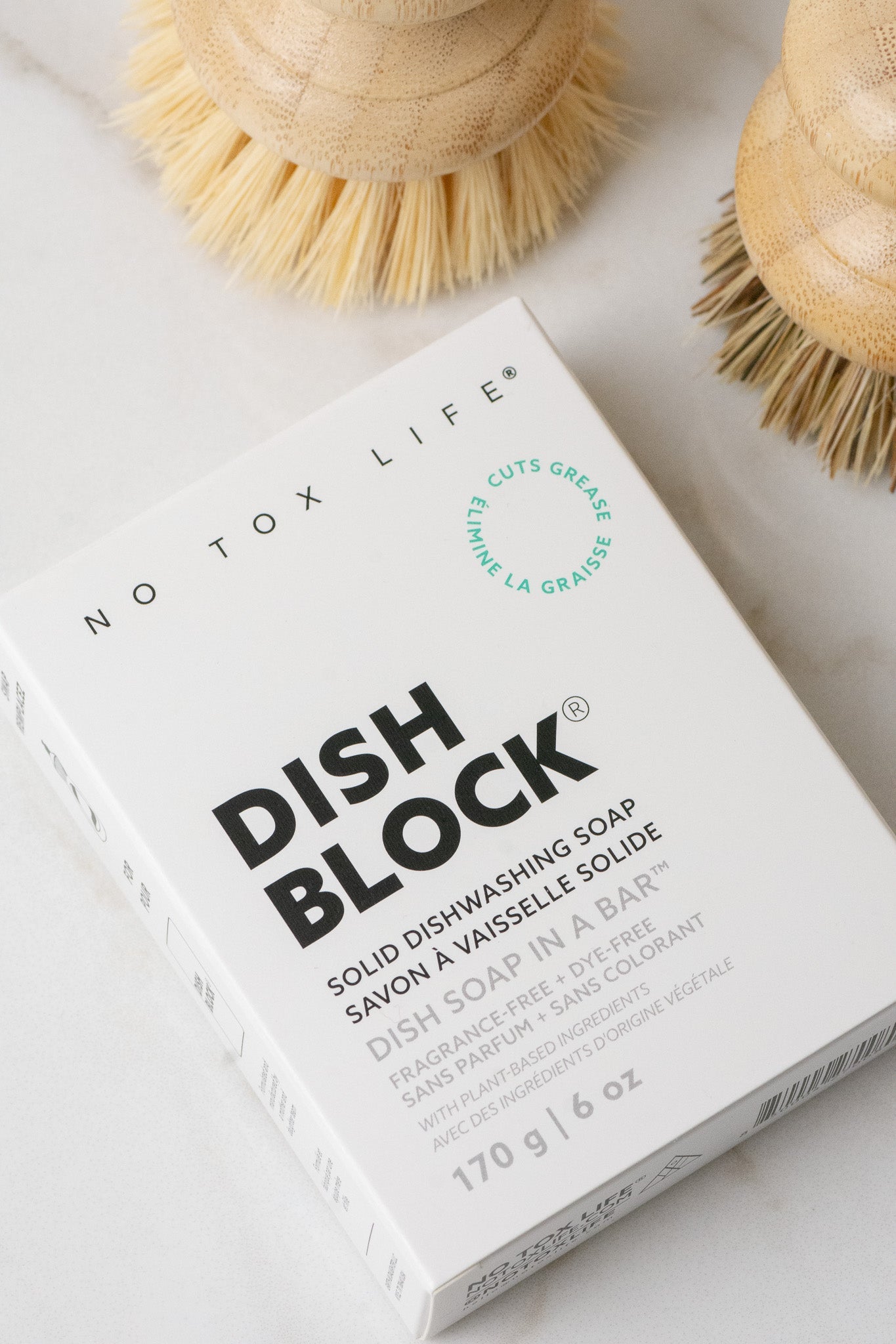 A white box labeled "DISH BLOCK® solid dish soap - 6 oz bar" by No Tox Life sits on a light surface with wooden brushes nearby. Minimal black text and green accents highlight its biodegradable, eco-friendly, grease-cutting formula.