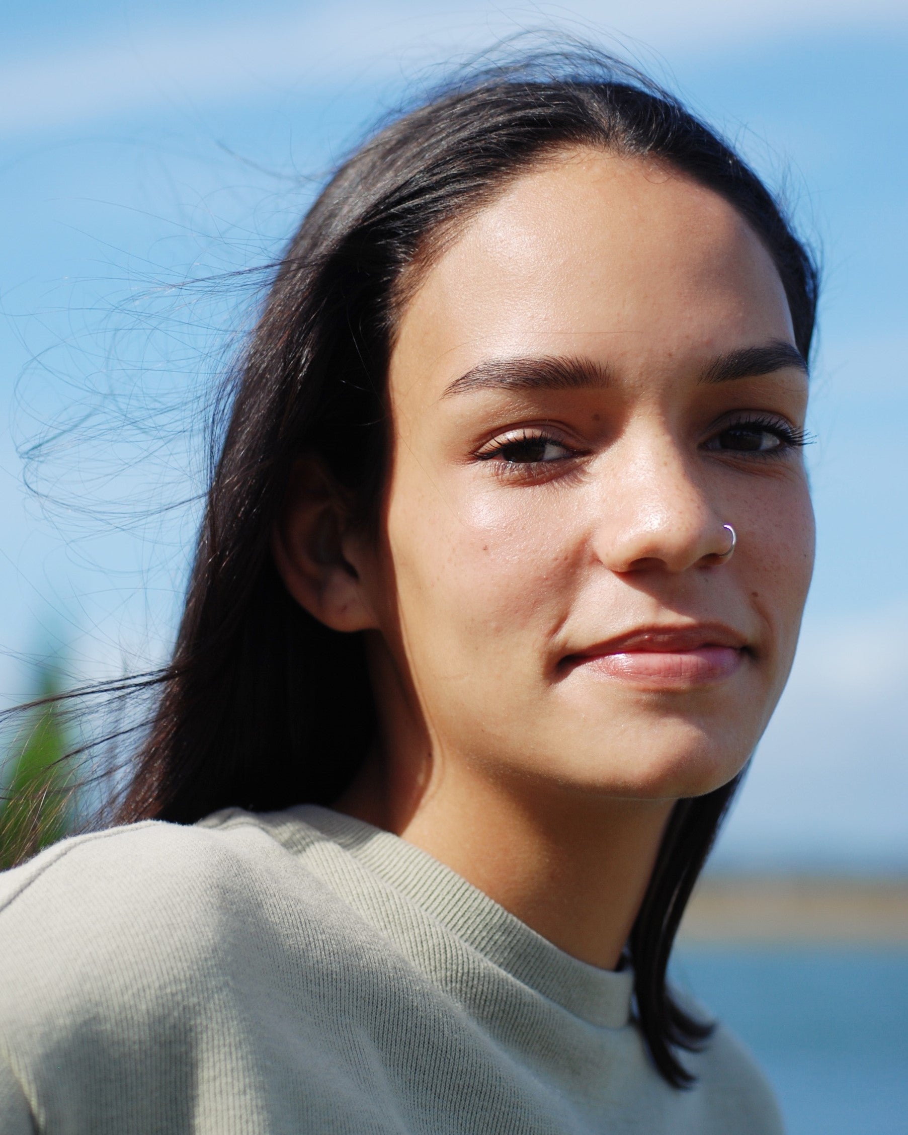 A young woman with straight dark hair and a nose ring smiles softly at the camera. She is wearing a light-colored top, and her natural brow color looks stunning under the sunlight, thanks to River Organics’ Tinted Vegan Eyebrow Wax | Dark. The background is blurred, showing hints of greenery and blue sky on a sunny day.