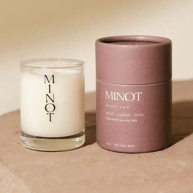 A Minot Luxury Non-toxic Soy Candle (3oz) sits beside a mauve cylindrical box labeled "MINOT daybreak" and "Rose Amber Oud," arranged on a neutral surface in soft, warm light for an inviting display.