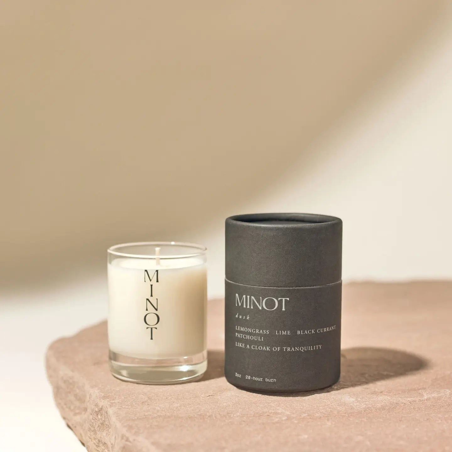 A Minot Luxury Non-toxic Soy Candle (3oz) in a minimalist clear glass holder sits beside a matching dark container, both on a textured stone surface, bathed in soft light and gentle shadows.