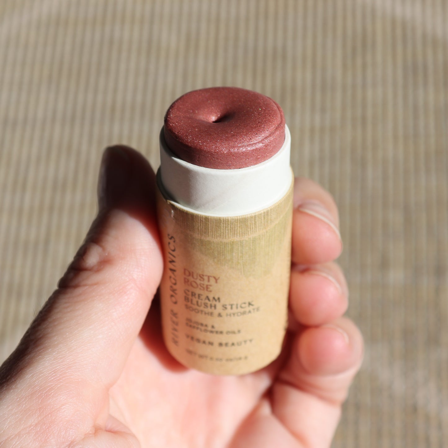 A hand presents the Creamy Vegan Blush Stick - Dusty Rose from River Organics against a textured backdrop. The cream stick features a muted rosy pink hue, reflecting Sustainable Beauty with its eco-friendly paper tube packaging.