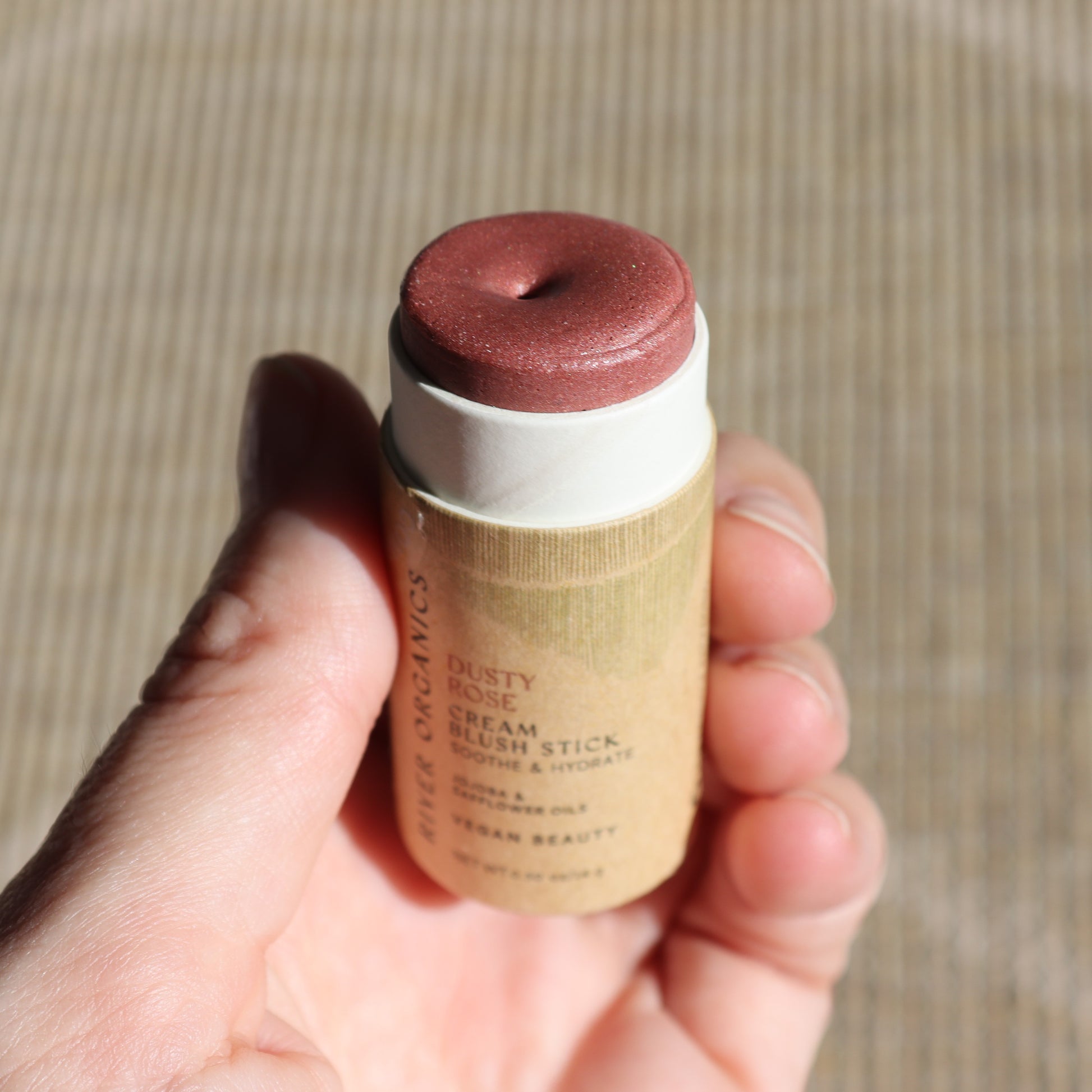A hand presents the Creamy Vegan Blush Stick - Dusty Rose from River Organics against a textured backdrop. The cream stick features a muted rosy pink hue, reflecting Sustainable Beauty with its eco-friendly paper tube packaging.