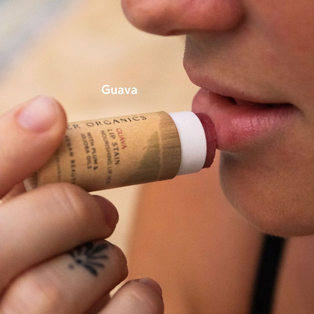 A person applies River Organics Vegan Lip Stains in "Guava" to their lips, holding the plastic-free tube near their mouth. A close-up showcases the eco-friendly lip color and a tattoo on their finger.