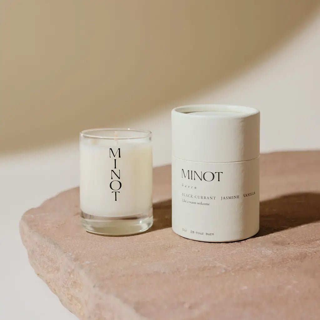 A 3oz Minot Luxury Non-toxic Soy Candle in a white glass holder sits beside a matching cylindrical box labeled "MINOT" and "BLACK CURRANT JASMINE VANILLA," all displayed on a light stone surface.
