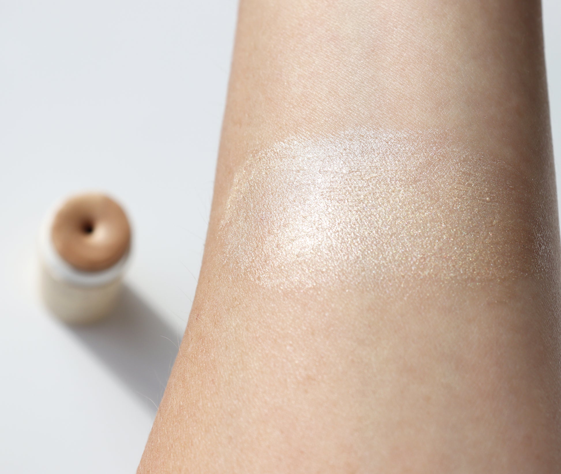 A close-up of a forearm displays a swatch of River Organics' Illumi Vegan Highlighter Stick in Golden Hue. In the background, a small, round container made from sustainable packaging holds the same shimmering shade and rests elegantly on a white surface.