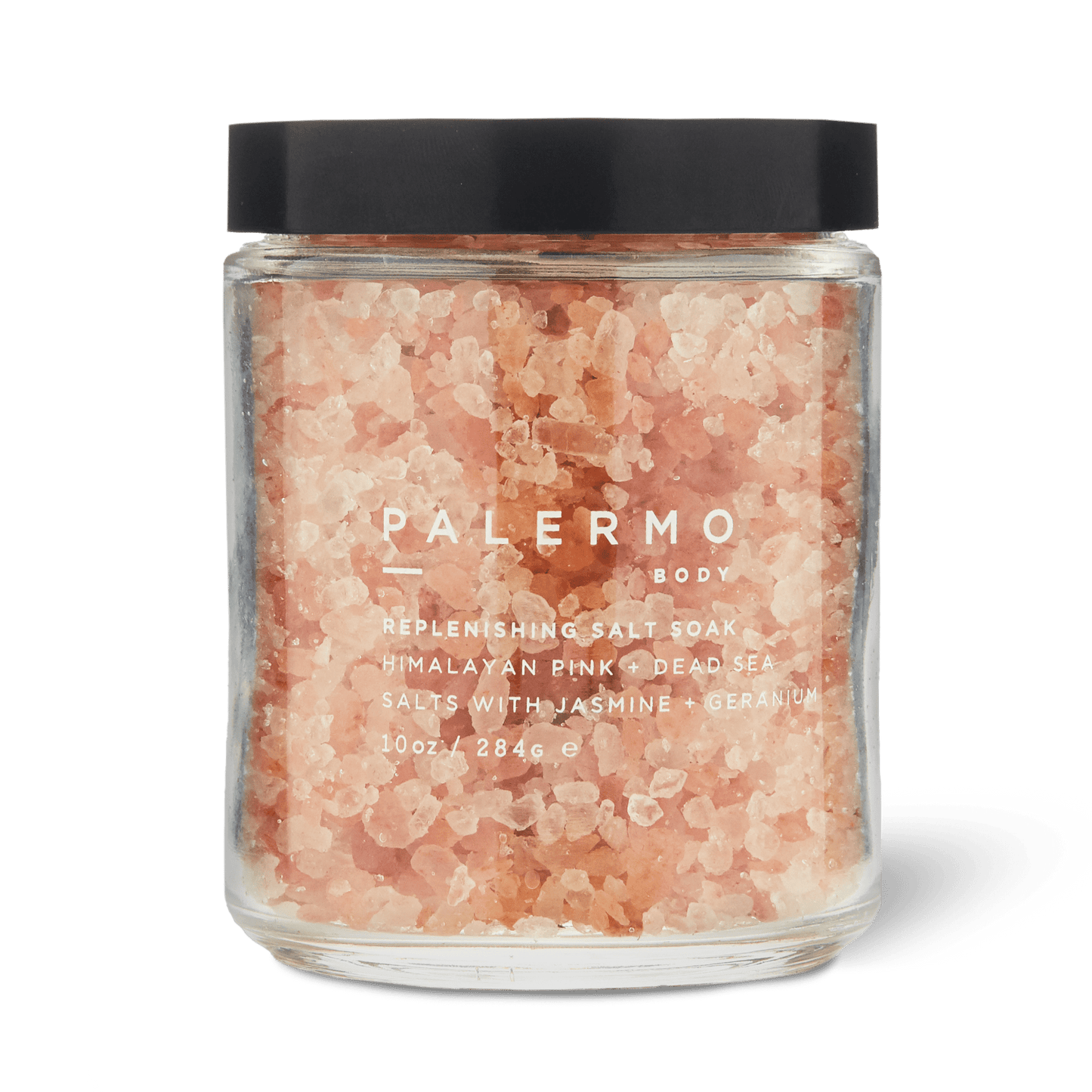 A clear glass jar with a black lid holds pink bath salts labeled “Palermo Body Replenishing Salt Soak.” This soothing jasmine and geranium bath soak combines Himalayan pink salt and Dead Sea salt for relaxation. (10 oz / 284.6 g).