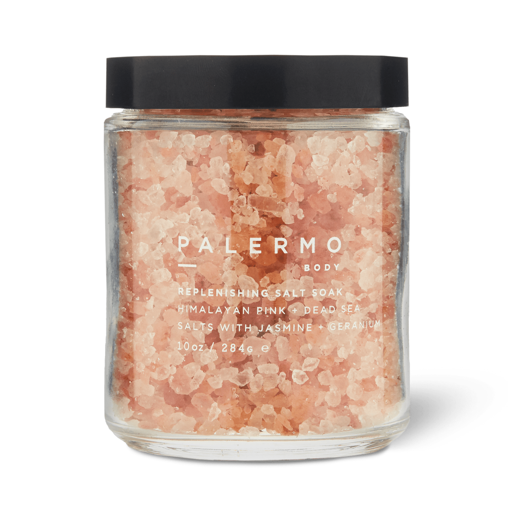 A clear glass jar with a black lid holds pink bath salts labeled “Palermo Body Replenishing Salt Soak.” This soothing jasmine and geranium bath soak combines Himalayan pink salt and Dead Sea salt for relaxation. (10 oz / 284.6 g).