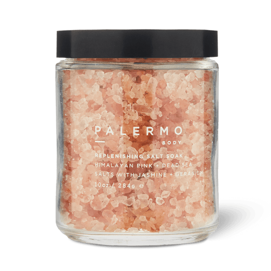 A clear glass jar with a black lid holds pink bath salts labeled “Palermo Body Replenishing Salt Soak.” This soothing jasmine and geranium bath soak combines Himalayan pink salt and Dead Sea salt for relaxation. (10 oz / 284.6 g).