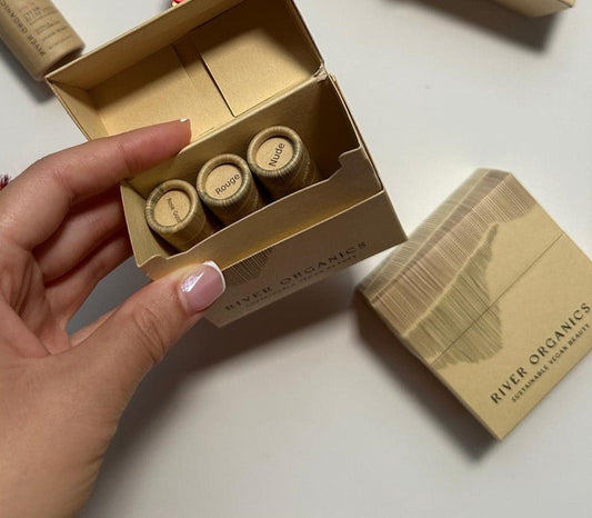 A hand holds an open River Organics box containing the Tinted Lip Balm Gift Set with three vegan lip balm tubes labeled "Rouge," "Nude," and one partially obscured, resting on a white surface with another box nearby.