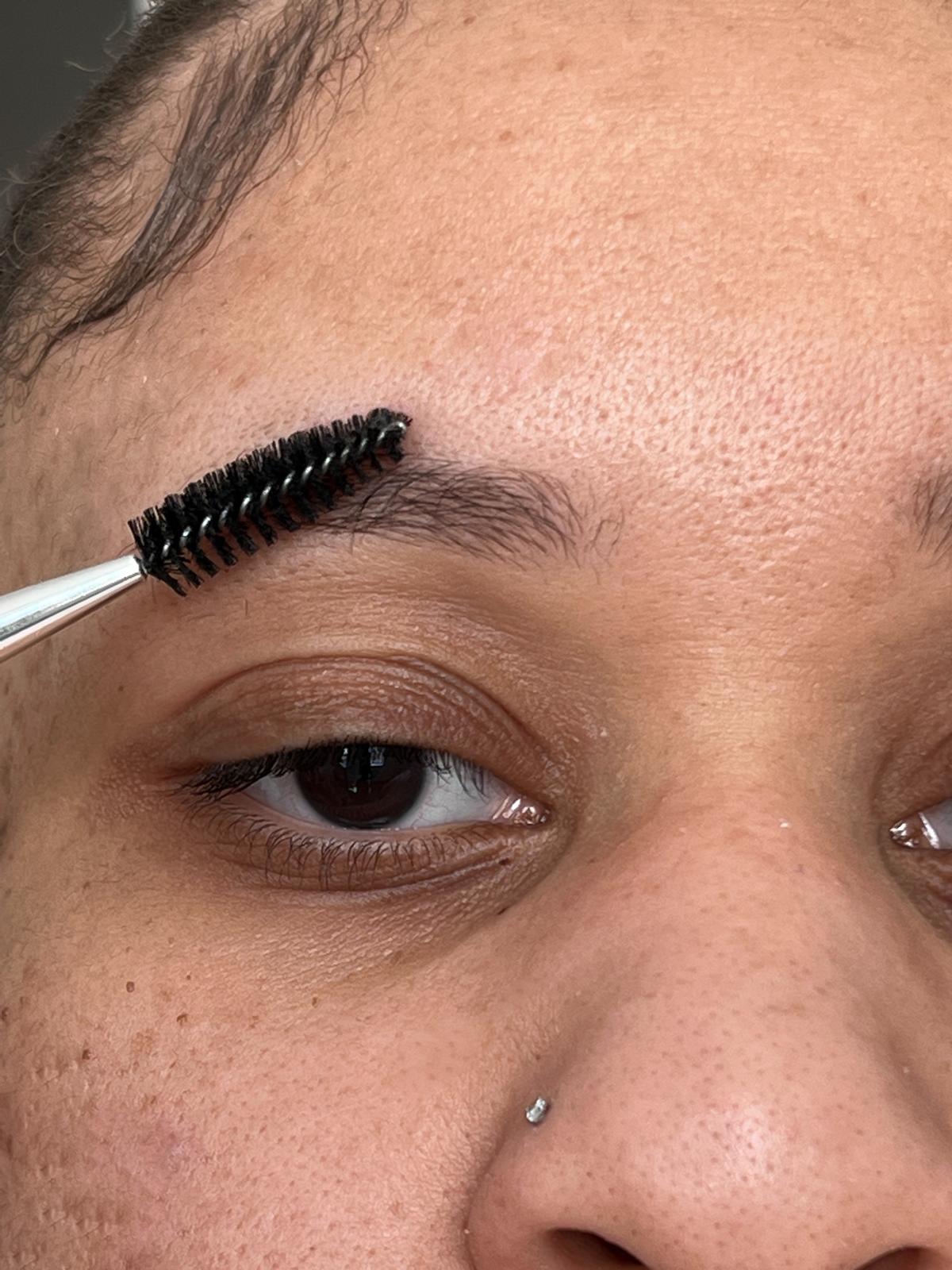 A close-up of someone expertly grooming their eyebrow with a spoolie brush and River Organics' Dark Vegan Shaping Eyebrow Wax, gently combing through brow hairs. A small nose piercing is seen on their left side against their visible skin.