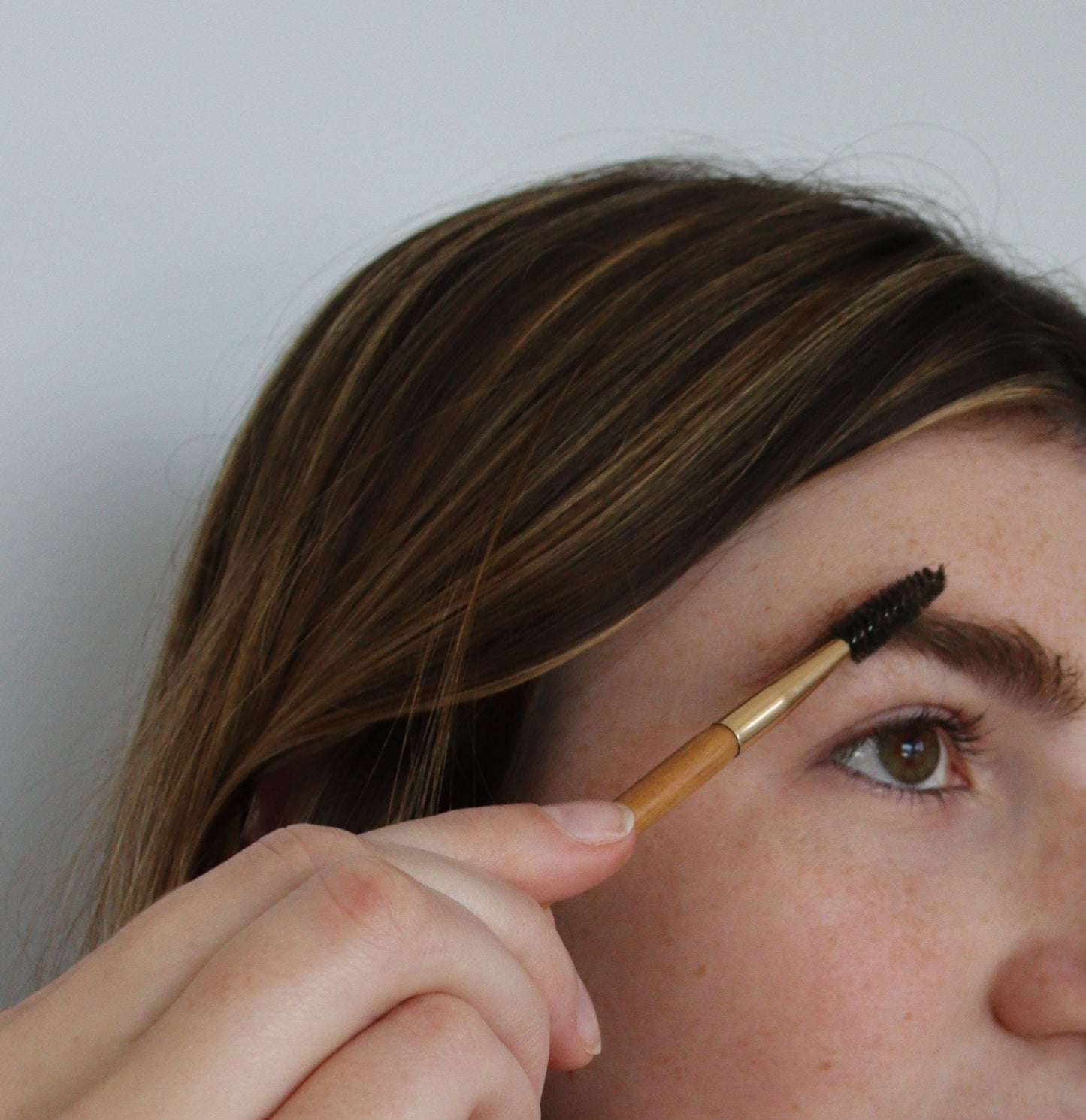 A person with brown hair is using a spoolie brush to groom their eyebrow with River Organics Skincare Tinted Vegan Eyebrow Wax | Medium Brown. The focus is on their hand holding the brush and their face, which is partially visible. The background is plain white.