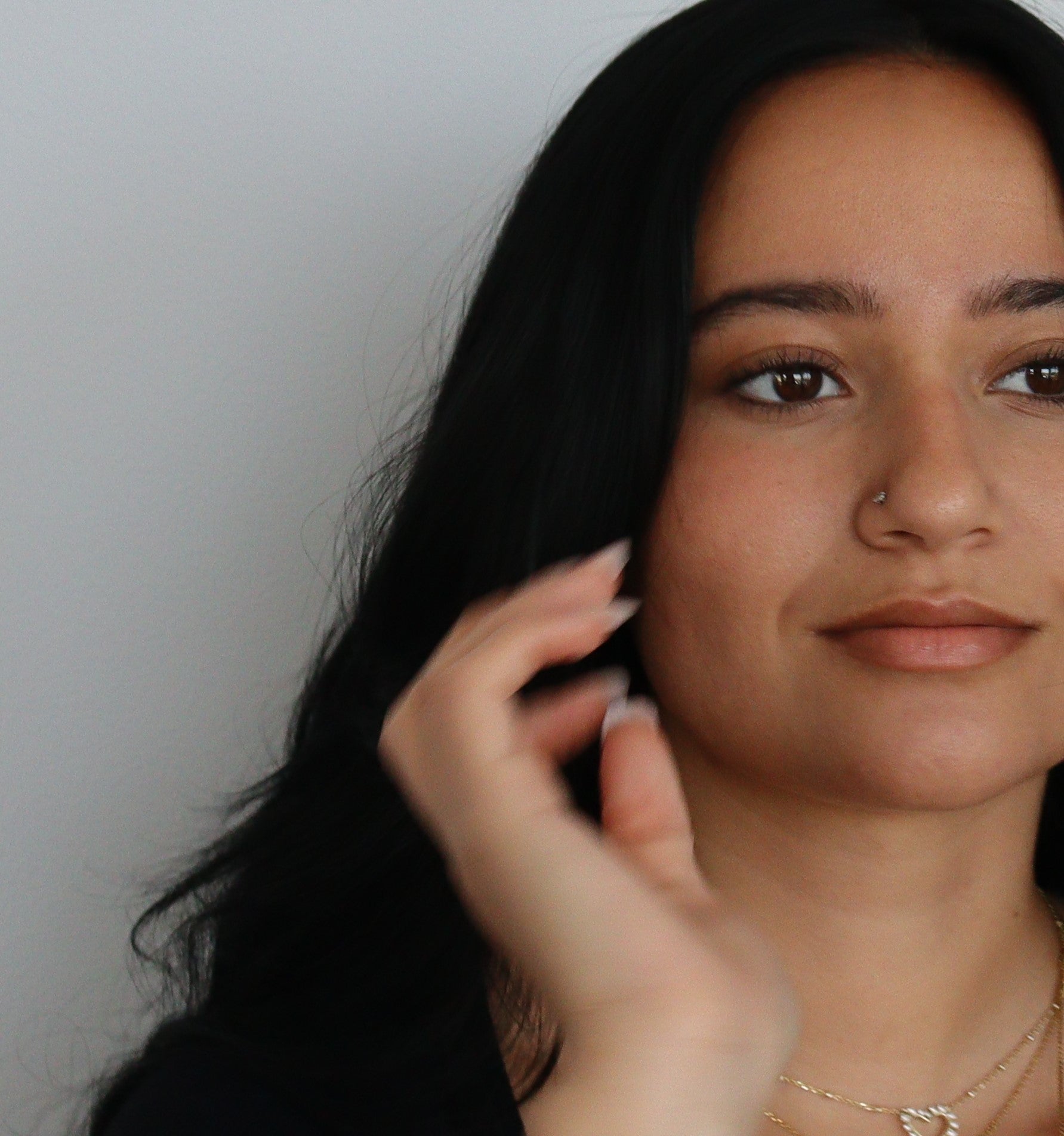 A person with long dark hair appears against a plain background, looking slightly to the right with a neutral expression. They wear a black top, nose ring, gold necklace, and have enhanced their complexion using River Organics' Shimmery Face and Body Bronzer Stick. One hand is raised near their face.