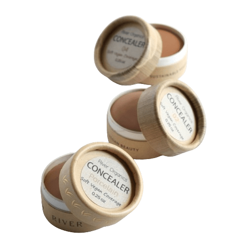 Three open containers of River Organics Zero-Waste Vegan Concealer with paper labels and wooden lids are displayed against a white background. These plant-based concealers in various shades feature certified organic oils, highlighting their commitment to sustainability.