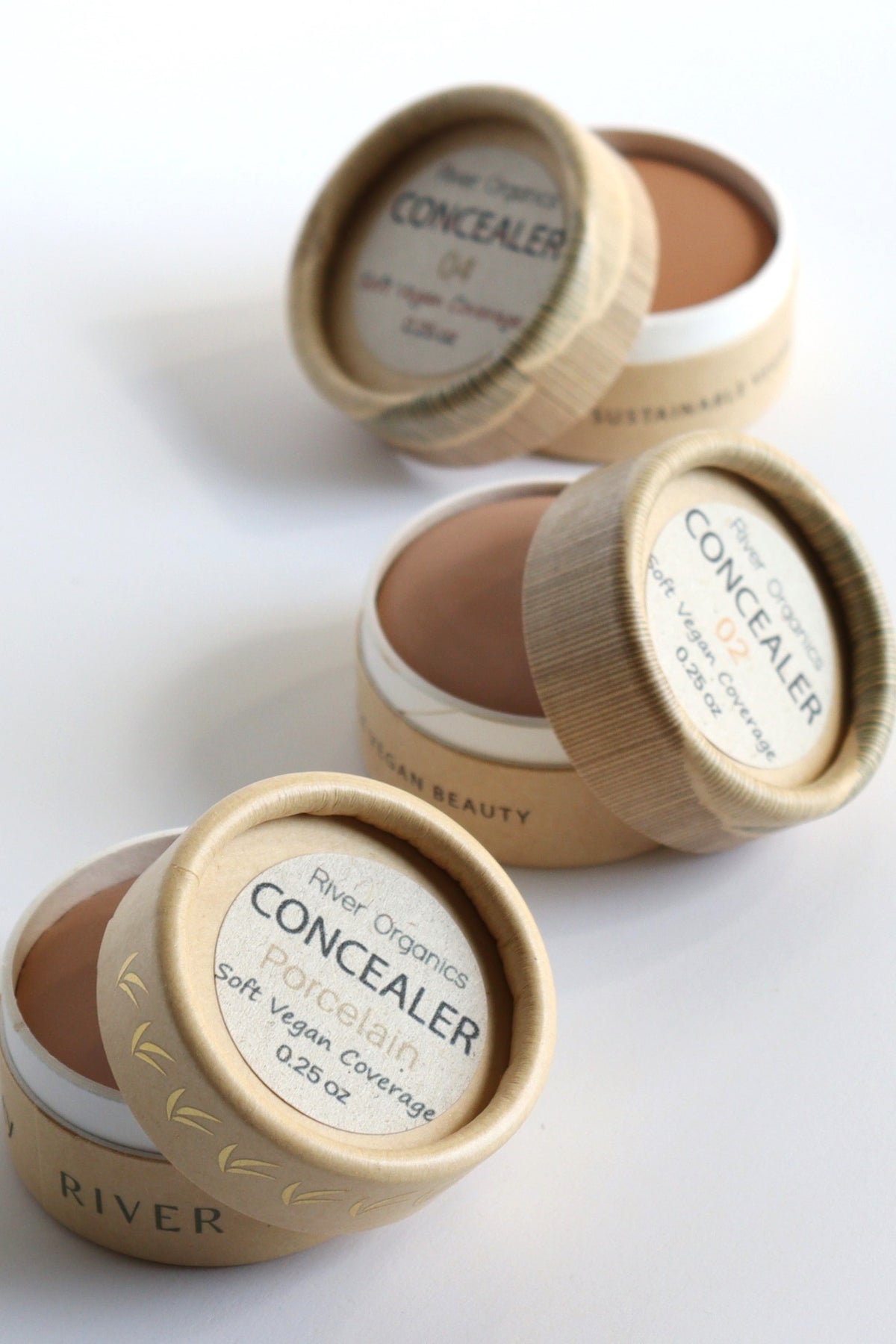 Four open containers of River Organics Imperfect Zero-Waste Vegan Concealer: Titanium dioxide-free, each in different shades with round cardboard lids, are arranged diagonally on a white surface. Enriched with organic plant oils and designed for zero-waste living, the labels emphasize its vegan, sustainable qualities and titanium dioxide-free formula.