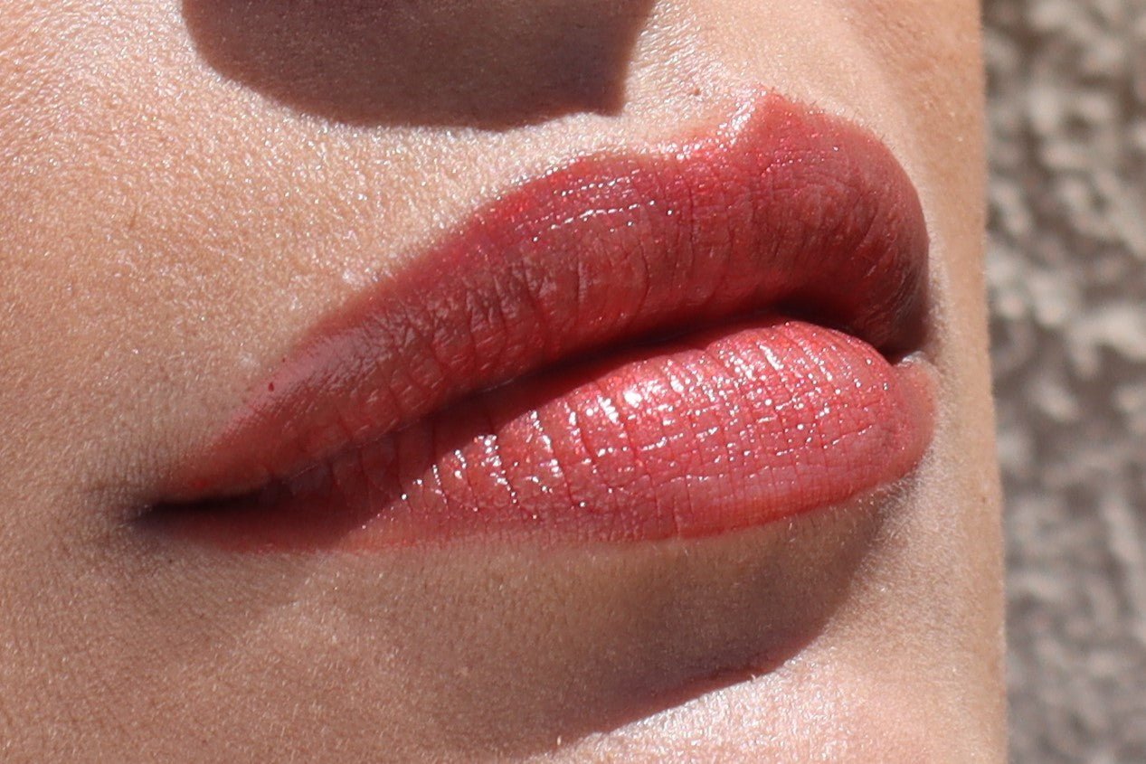 Close-up image of a person's lips wearing glossy red lipstick. The lips are slightly parted, and the light reflects off the glossy texture, highlighting the rich color and smooth application. This Pomegranate Vegan Lip Stain by River Organics ensures both vibrant shades and moisturizing lip care. The background is out of focus and appears neutral.