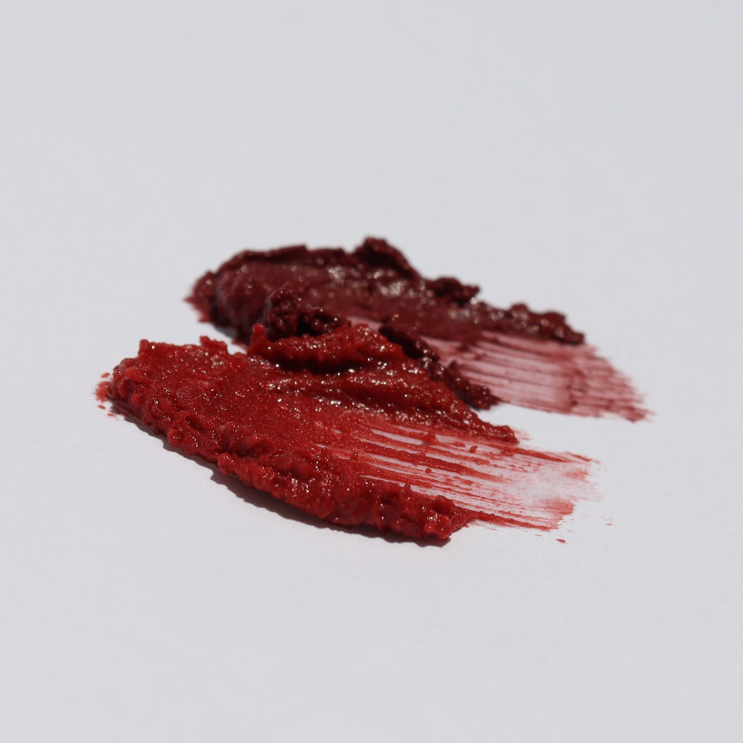 Two smears of River Organics' Lip Stain Set in "Fig" and "Pomegranate" on a white surface showcase the thick, textured vegan formula with varying shades from dark to slightly lighter red.