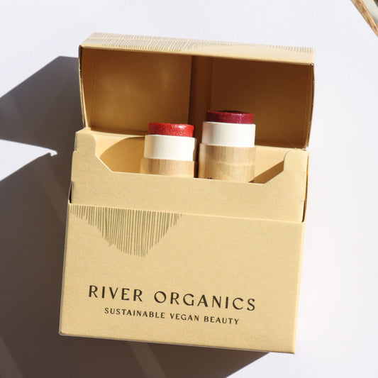 A white surface holds a cardboard box labeled "River Organics," featuring a Lip Stain Set with "Fig" and "Pomegranate." Inside are two cylindrical tubes filled with red vegan lip balms, infused with organic shea butter, casting soft shadows.