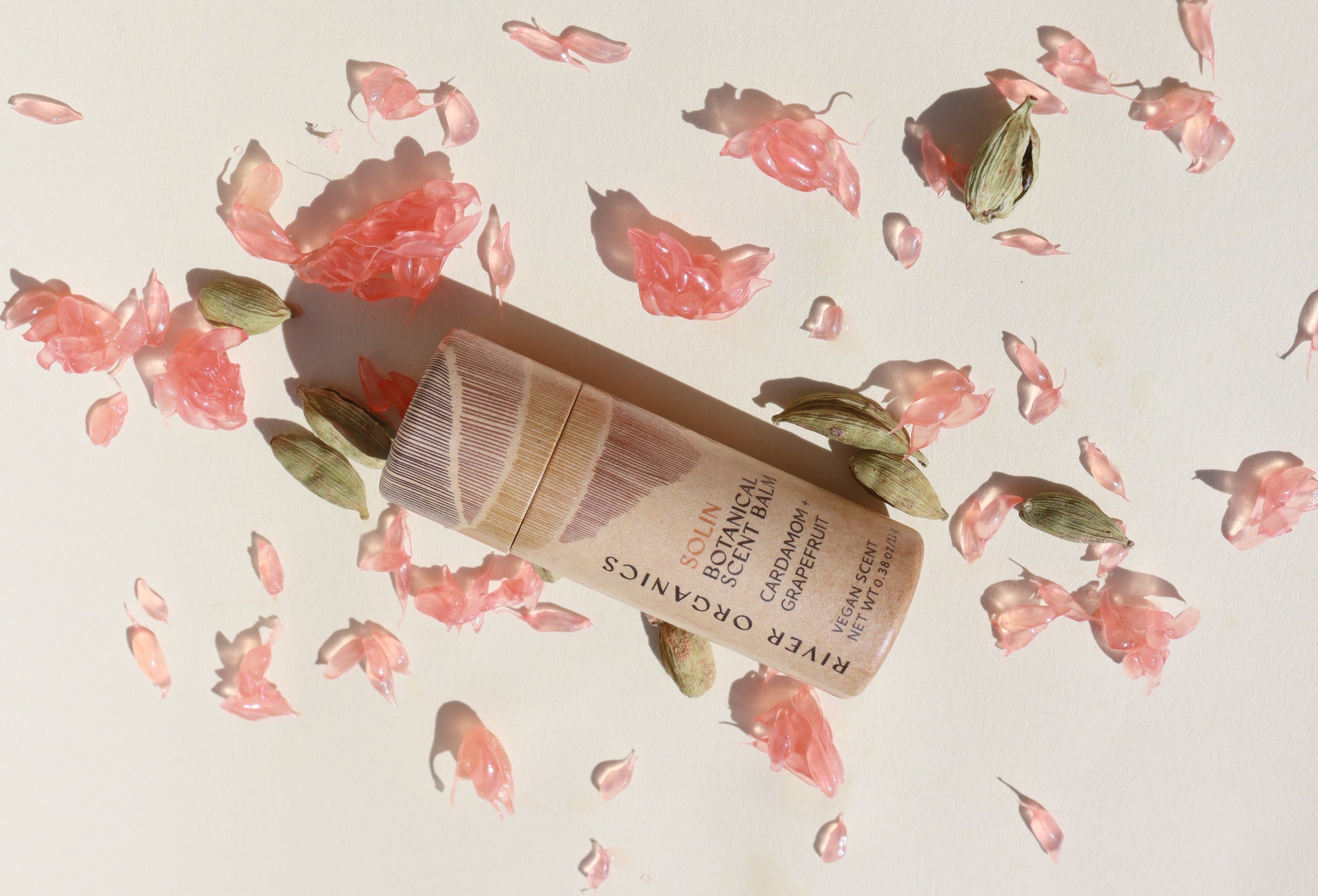 The Solin: Cardamom + Grapefruit Botanical Scent Balm by River Organics rests on a light surface, surrounded by dried pink petals and green cardamom pods, highlighting its gentle, natural fragrance.