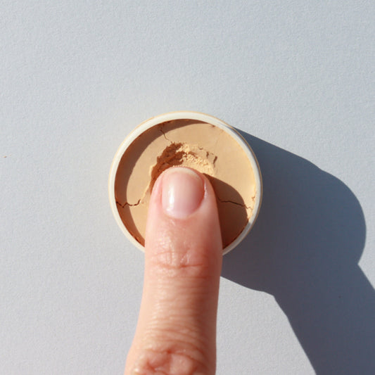 A finger presses into a small circular container of River Organics Zero-Waste Vegan Concealer - Titanium Dioxide-Free.
