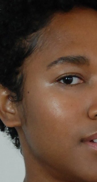 Close-up of the left side of a person’s face with dark skin and short curly hair, their eye, cheek, and lips glowing warmly from River Organics’ Illumi Cream Vegan Highlighter Stick, all enhanced by soft lighting.