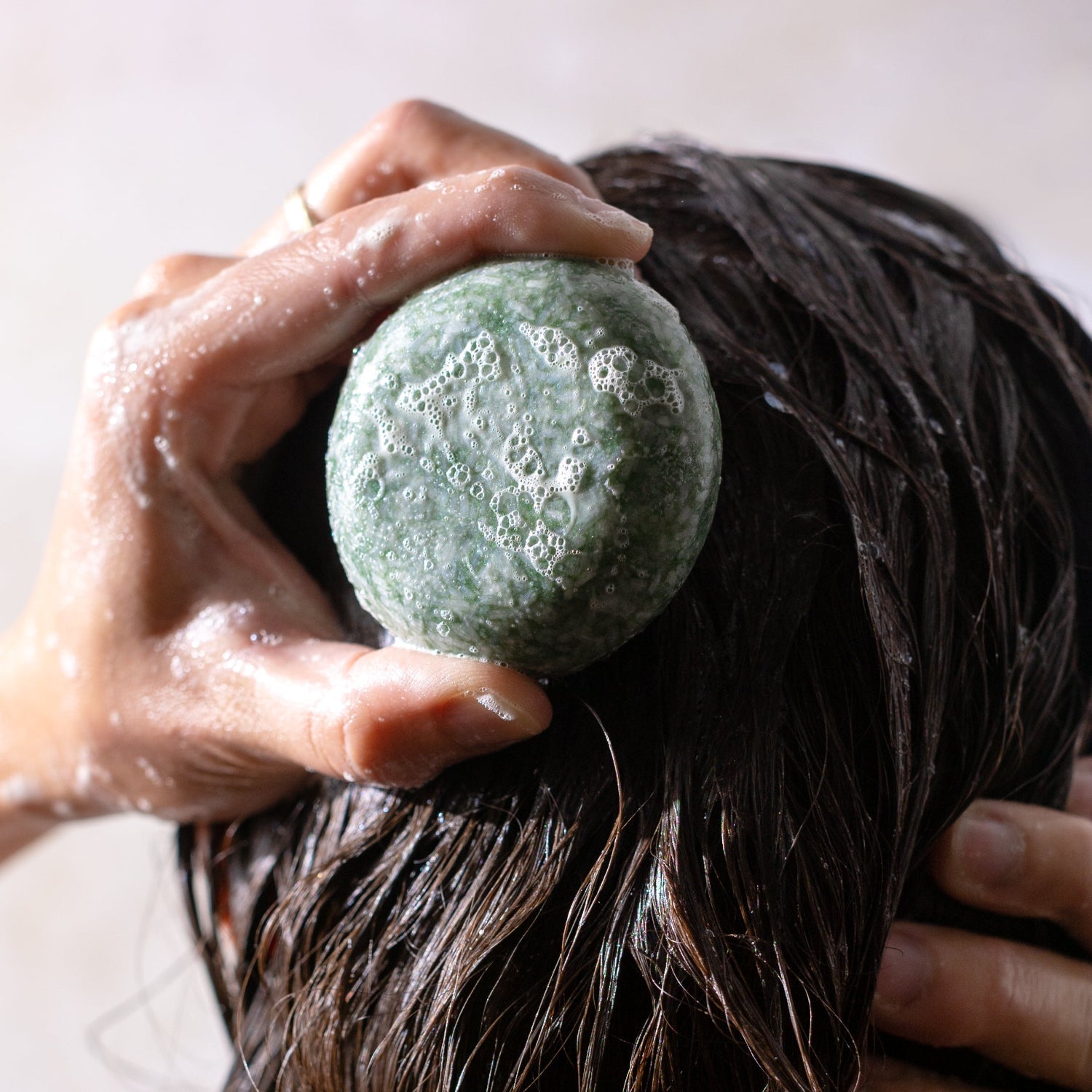 A hand holds the ZeroWasteStore.com Shampoo Bar, lathering the green, round bar onto wet, dark hair to create bubbles and foam.