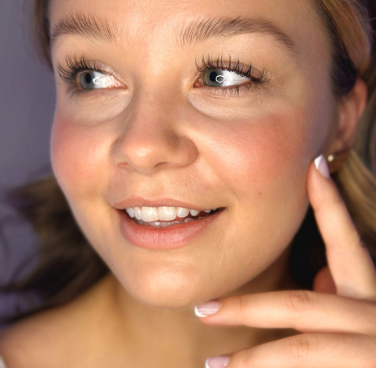 A woman with wavy brown hair smiles gently, revealing her teeth. She wears natural makeup highlighted by a light pink shade of the River Organics Natural Cream Blush Stick, and her manicured nails lightly touch her face. The background is softly blurred.