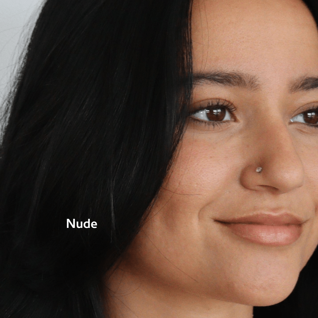 A person with long dark hair and a nose piercing smiles softly, wearing River Organics Tinted Vegan Lip Balm. The word “Nude” appears in their hair, while only part of their face is visible against a light background.