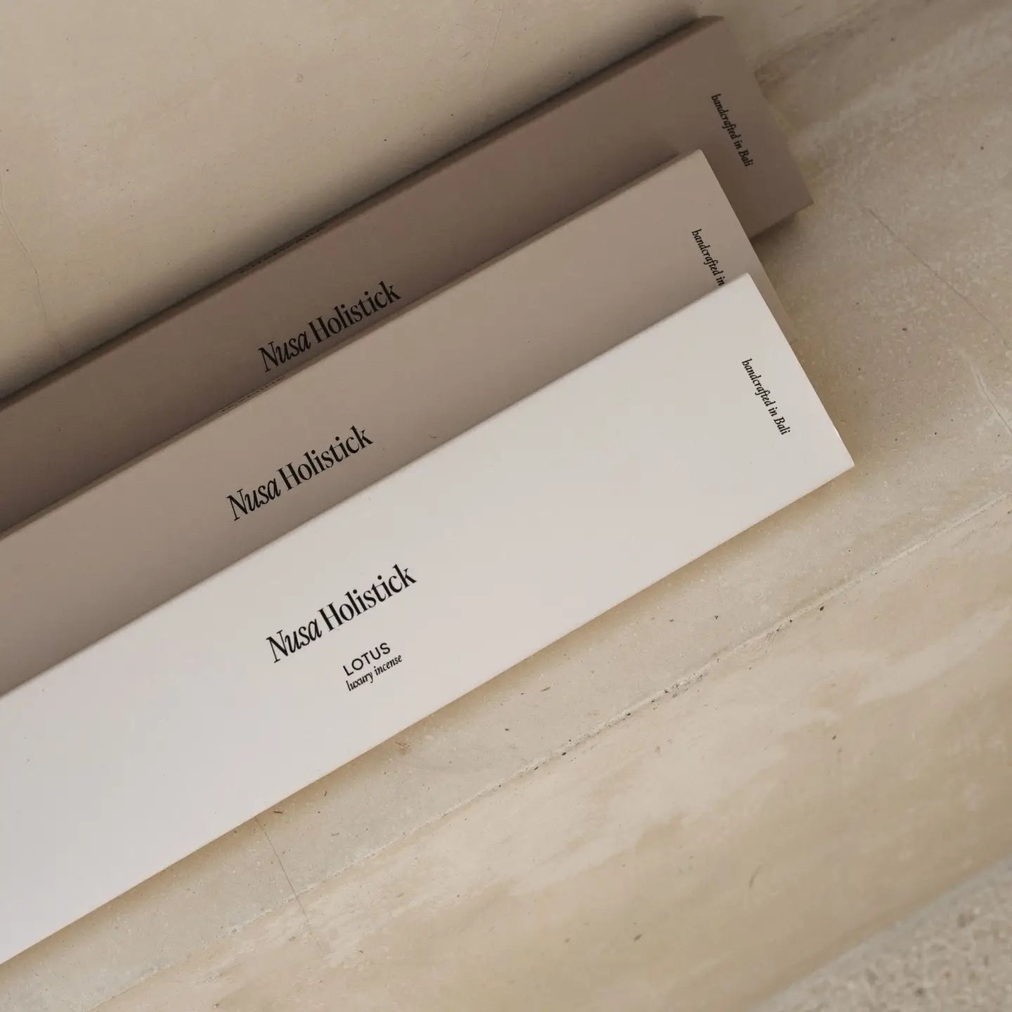 Three neutral-toned boxes labeled "Nusa Holistick" are arranged diagonally on a light stone surface. The front box displays "Essential Oil Incense – Jasmine, Lotus, Champaka," reflecting Bali-inspired handcrafted scents.