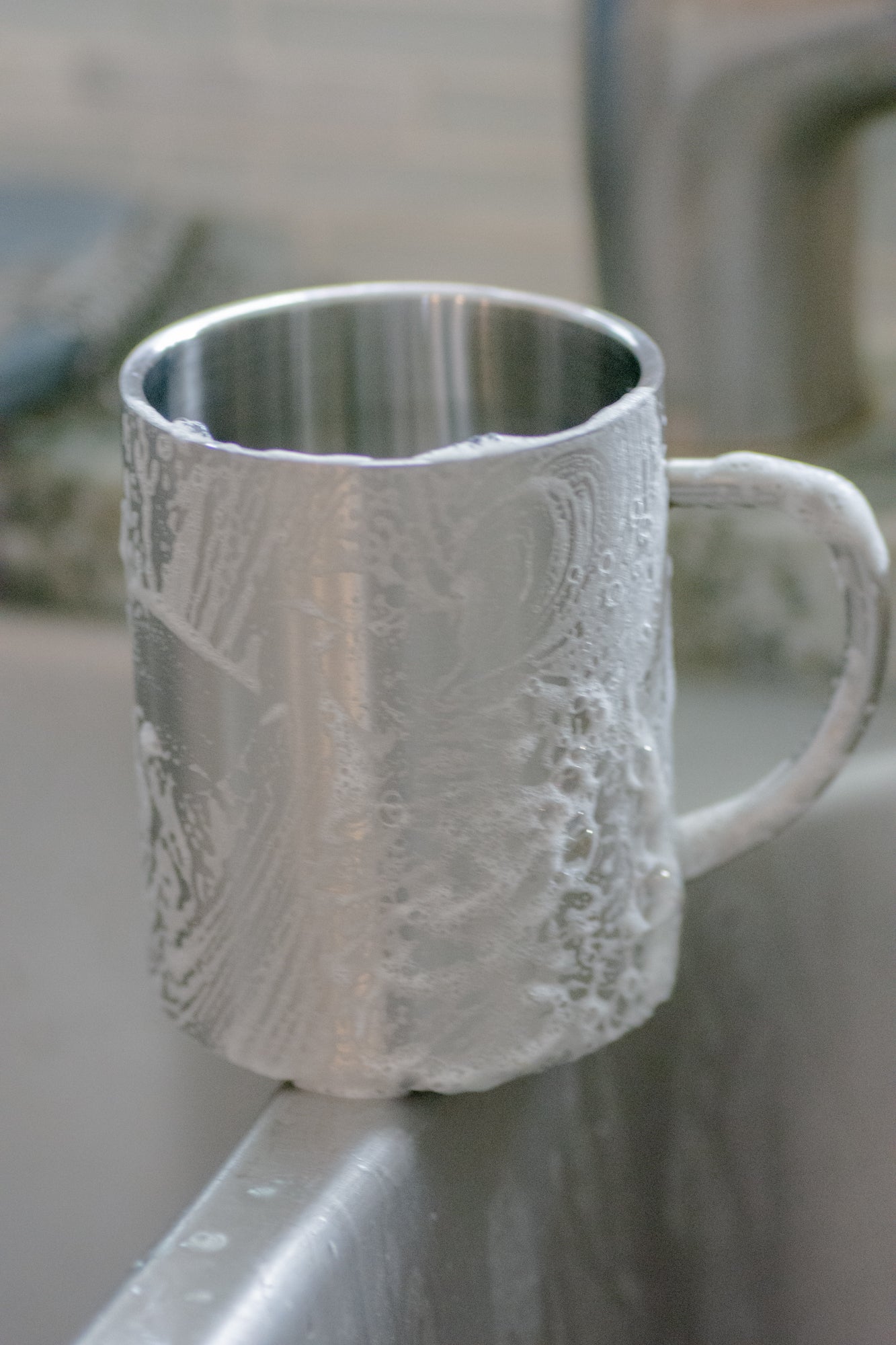 A metal mug covered in suds rests on the sink’s edge, with bubbles created by No Tox Life DISH BLOCK® solid dish soap. The background is softly out of focus.