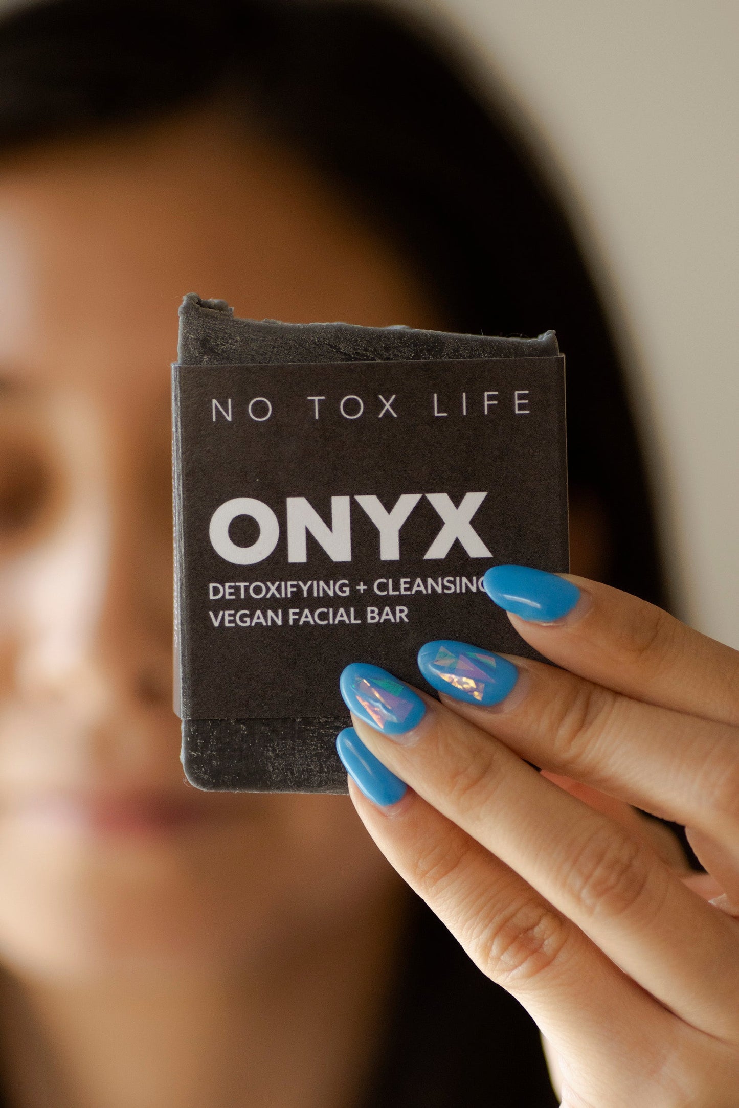 A person with blue decorated nails holds up a NO TOX LIFE SOLIDSILK® ONYX Facial Cleansing Bar, while their face appears blurred in the background.