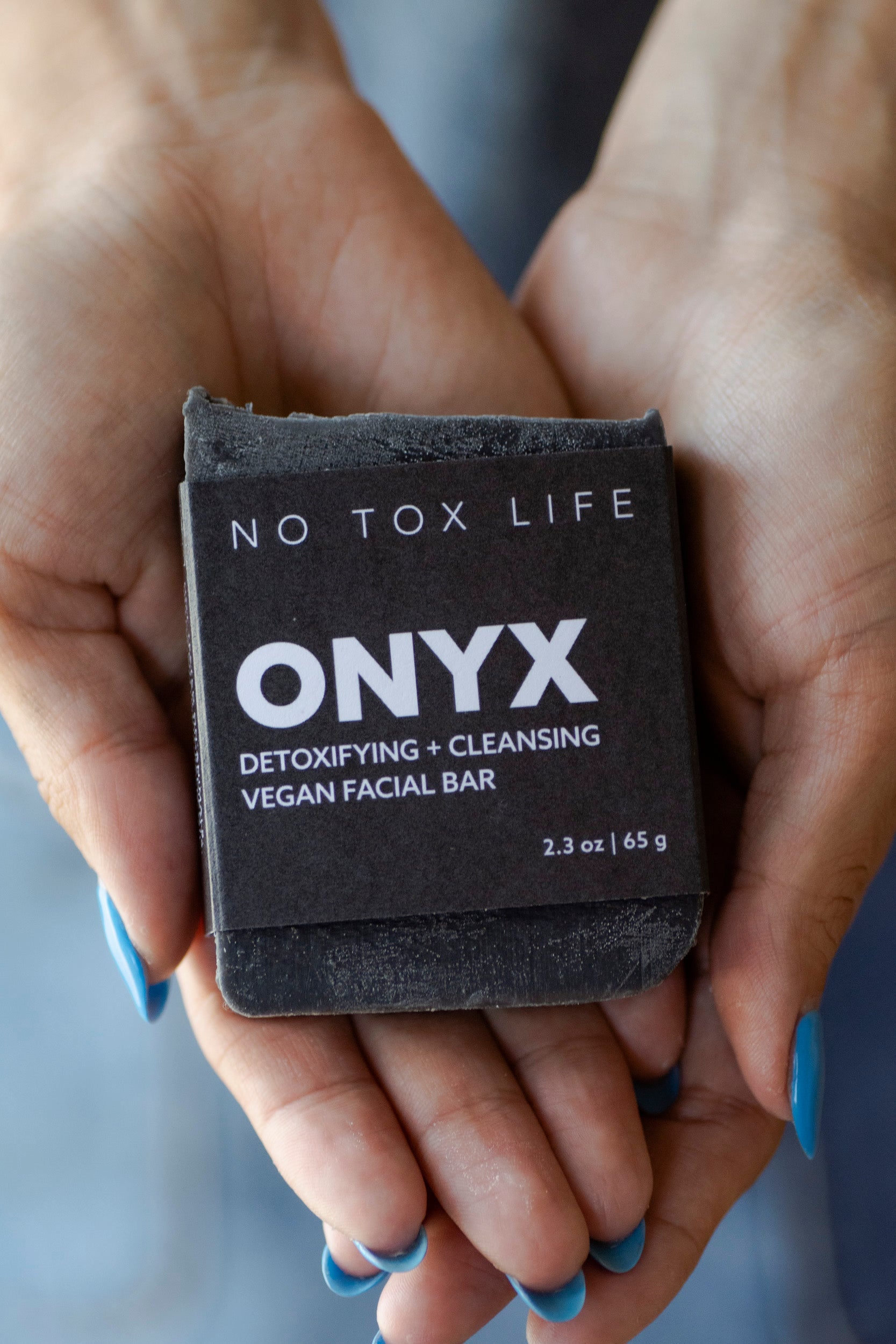 A person with blue-painted nails holds the NO TOX LIFE SOLIDSILK® ONYX Facial Cleansing Bar, a vegan cleanser with activated charcoal, in their cupped hands.