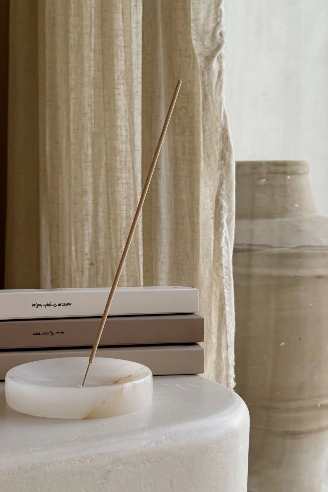 A lit Nusa Holistick Essential Oil Incense stick (Jasmine, Lotus, Champaka) in a white holder sits on a table beside neutral books, with beige curtains and a large ceramic vase creating a calm, minimalist scene.