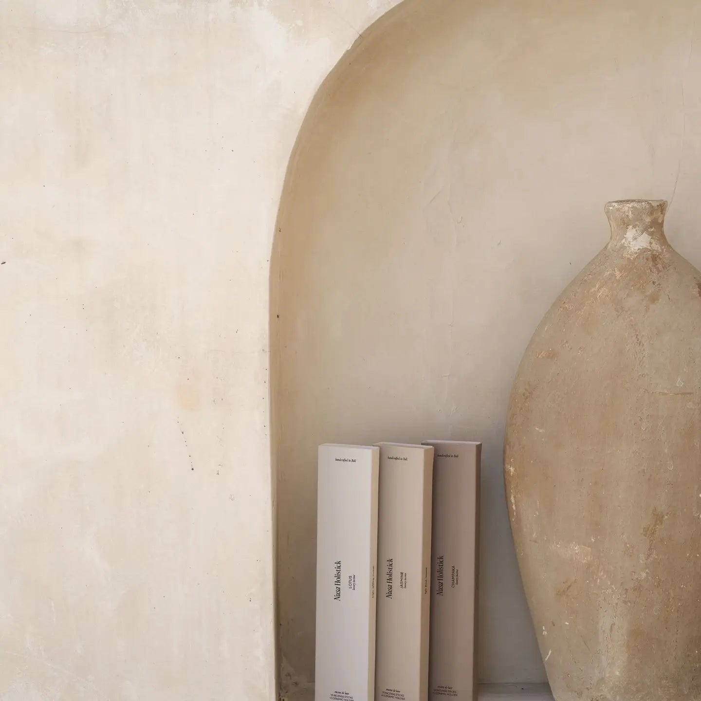 A large, rustic beige vase stands in a curved wall niche beside three slim boxes of Nusa Holistick Essential Oil Incense—Jasmine, Lotus, Champaka—set against a textured off-white wall.